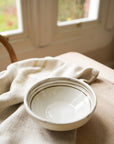 A hand painted ceramic bowl with breton stripes, placed on a linen cloth near a window.
