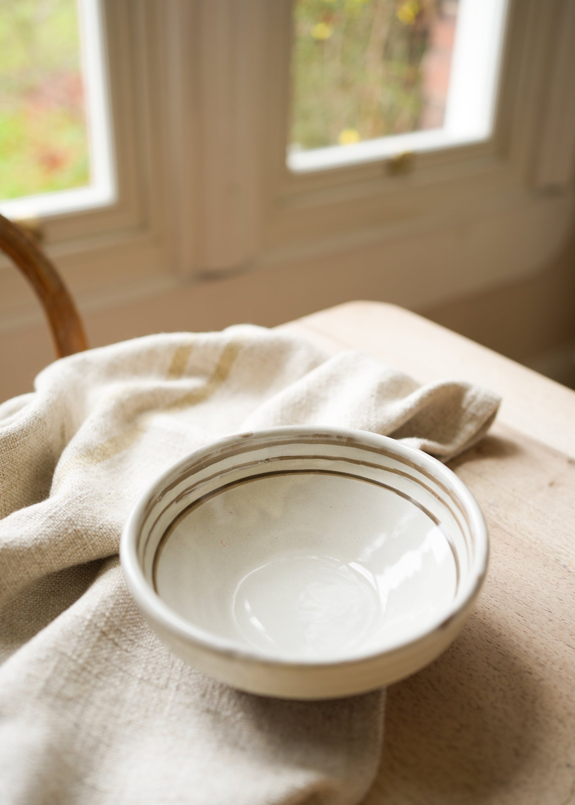 A hand painted ceramic bowl with breton stripes, placed on a linen cloth near a window.